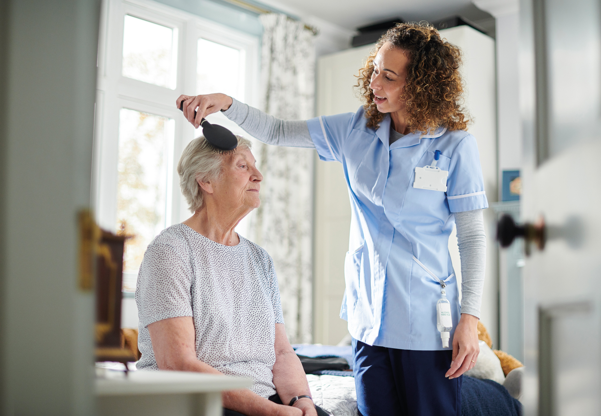 An older woman sitting down having her hair brushed by a female carer within a homely environment