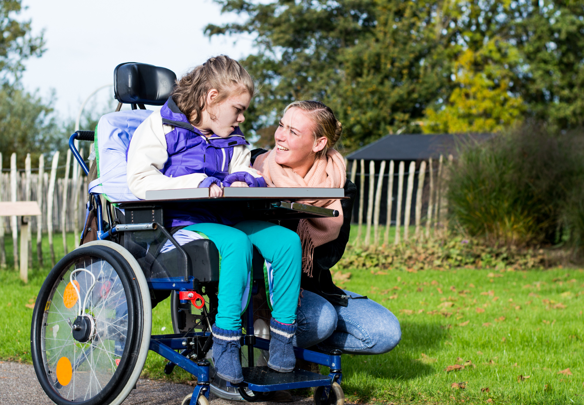 A young disabled girl in a wheelchair with a female carer beside her within an outside environment