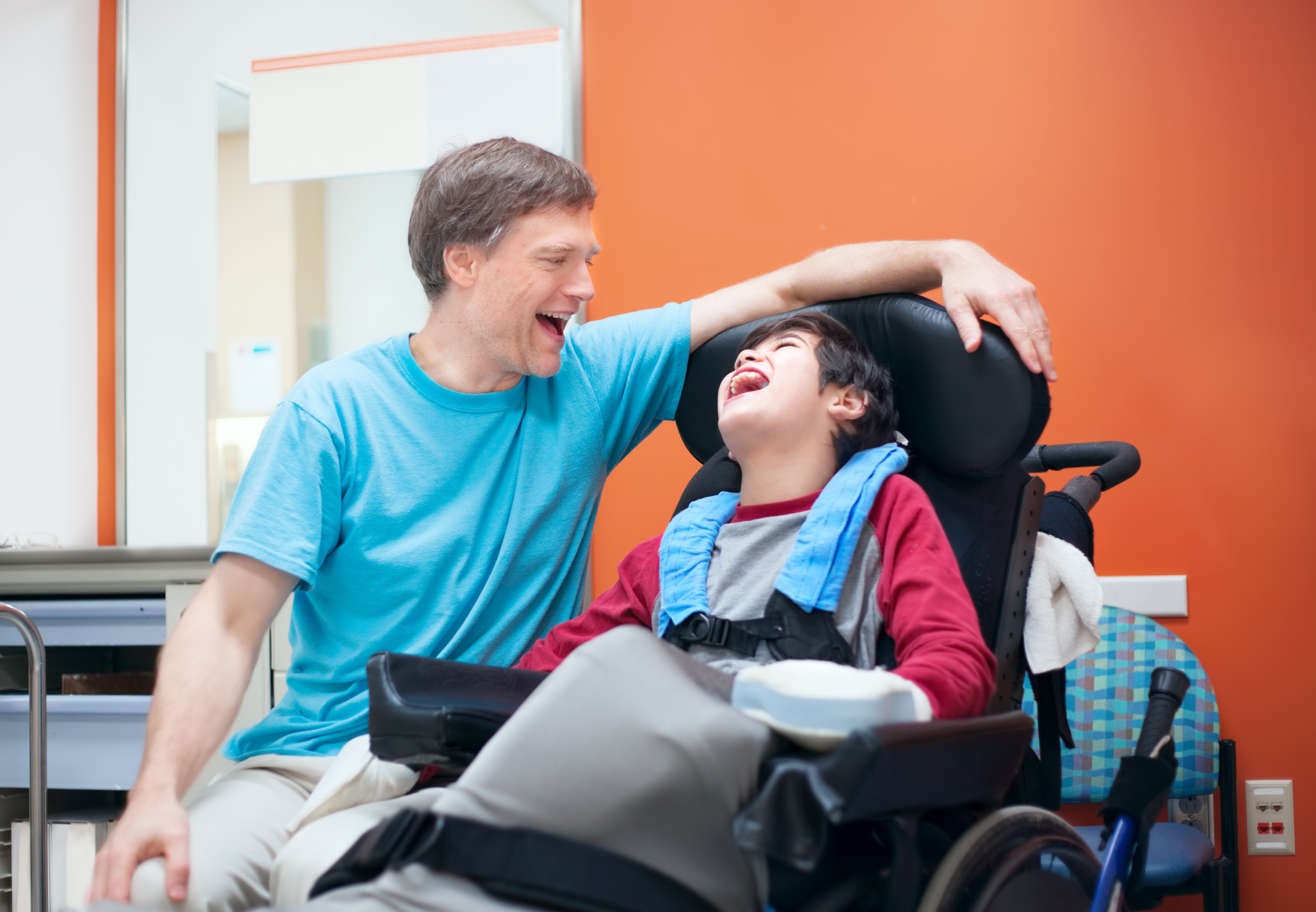 A young disabled boy in a wheelchair laughing with his male carer beside him