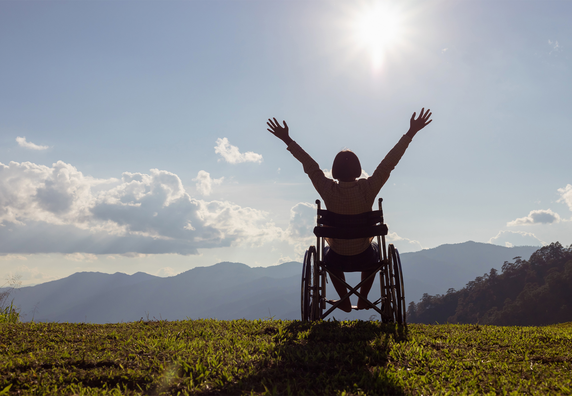 A disabled woman in a wheelchair with her hands in the air happily on a hill, surrounded by a landscape view
