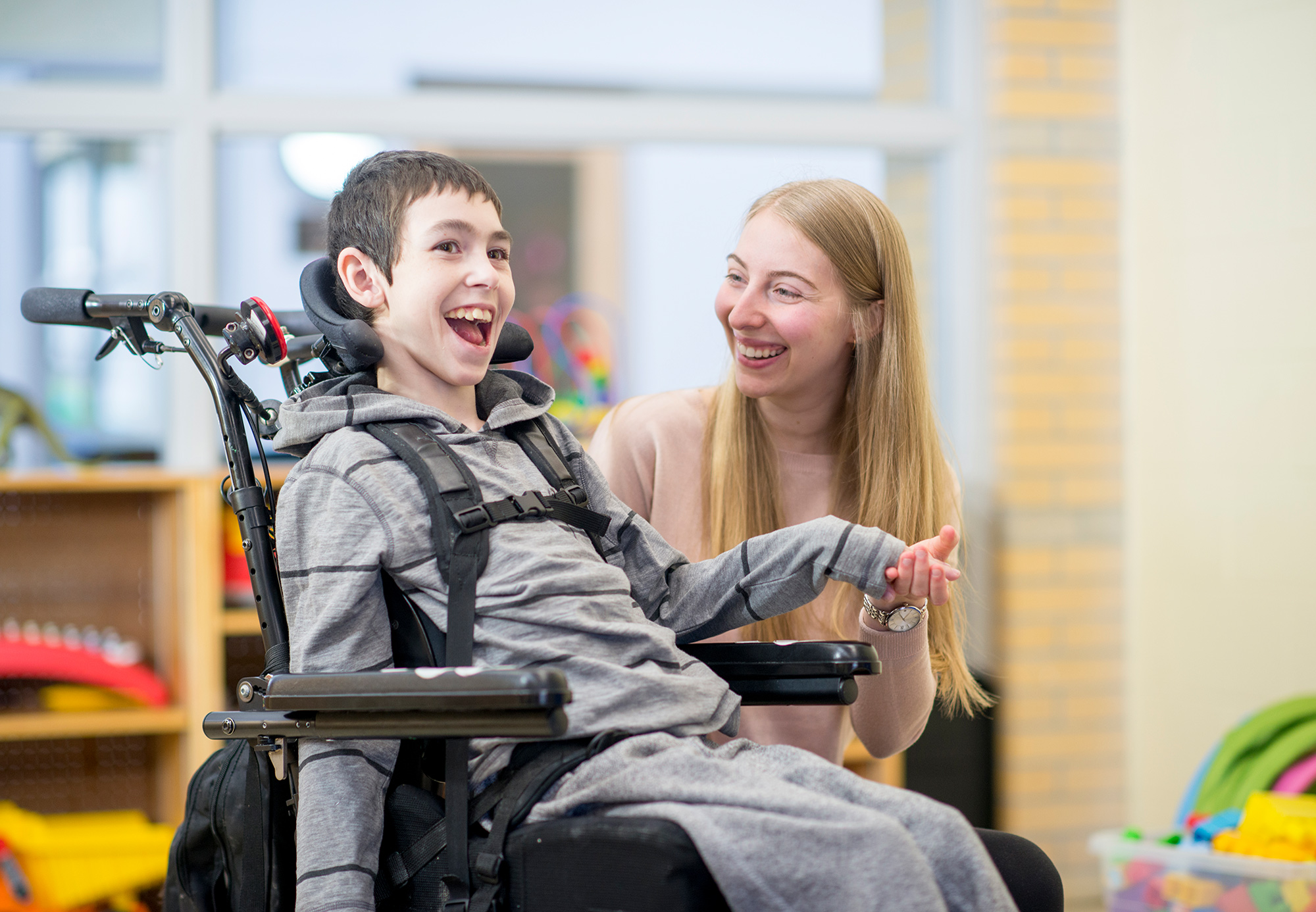 A young, happy disabled boy in a wheelchair next to a carer within a playroom environment