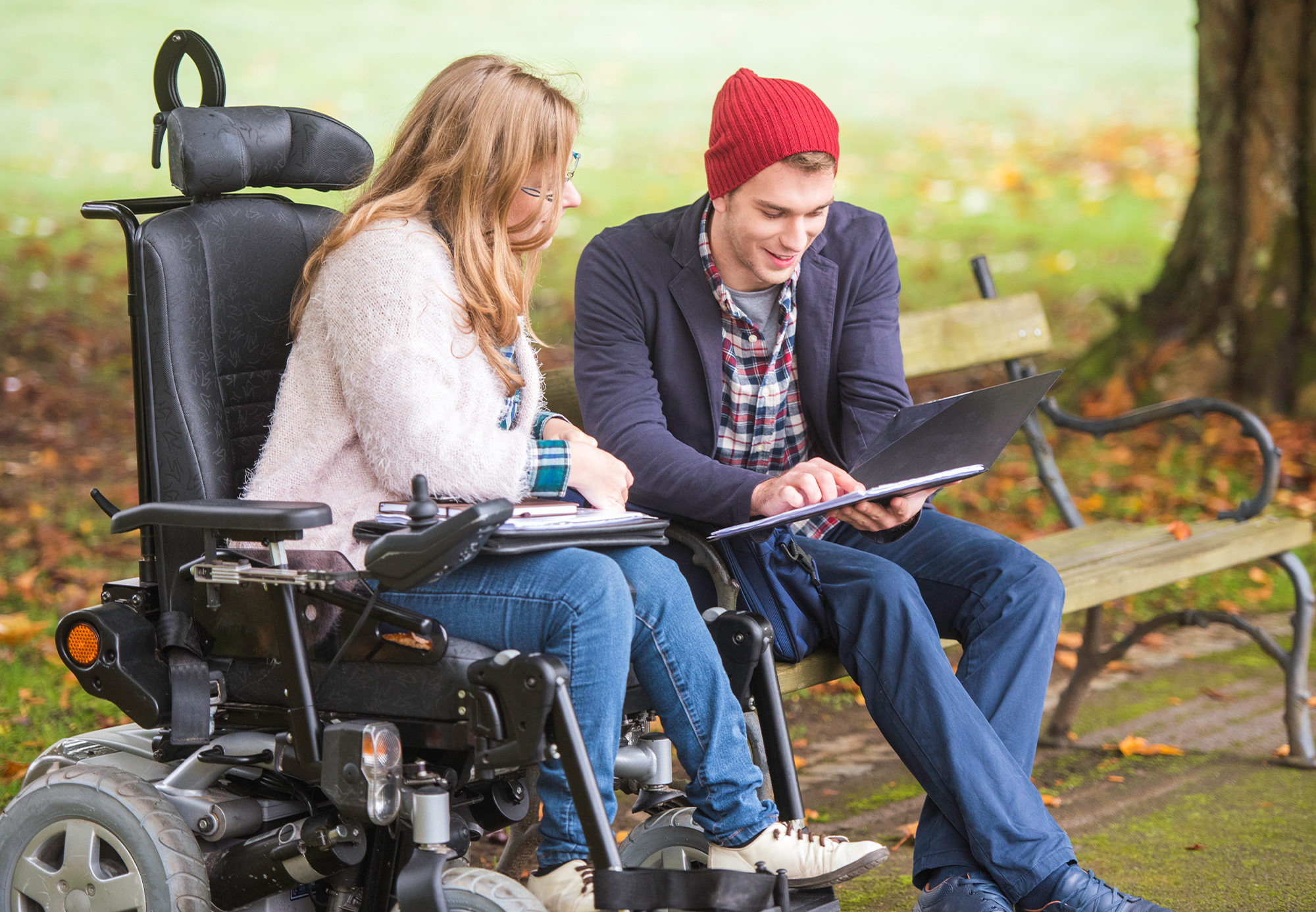 A disabled woman in a wheelchair beside her male carer looking at a filing, within a park